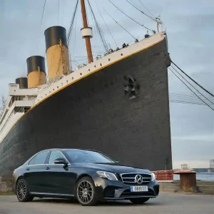 Black Mercedes chauffeur car parked beside a large ocean liner with yellow funnels at the dockside