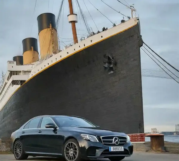 Black Mercedes chauffeur car parked beside a large ocean liner with yellow funnels at the dockside
